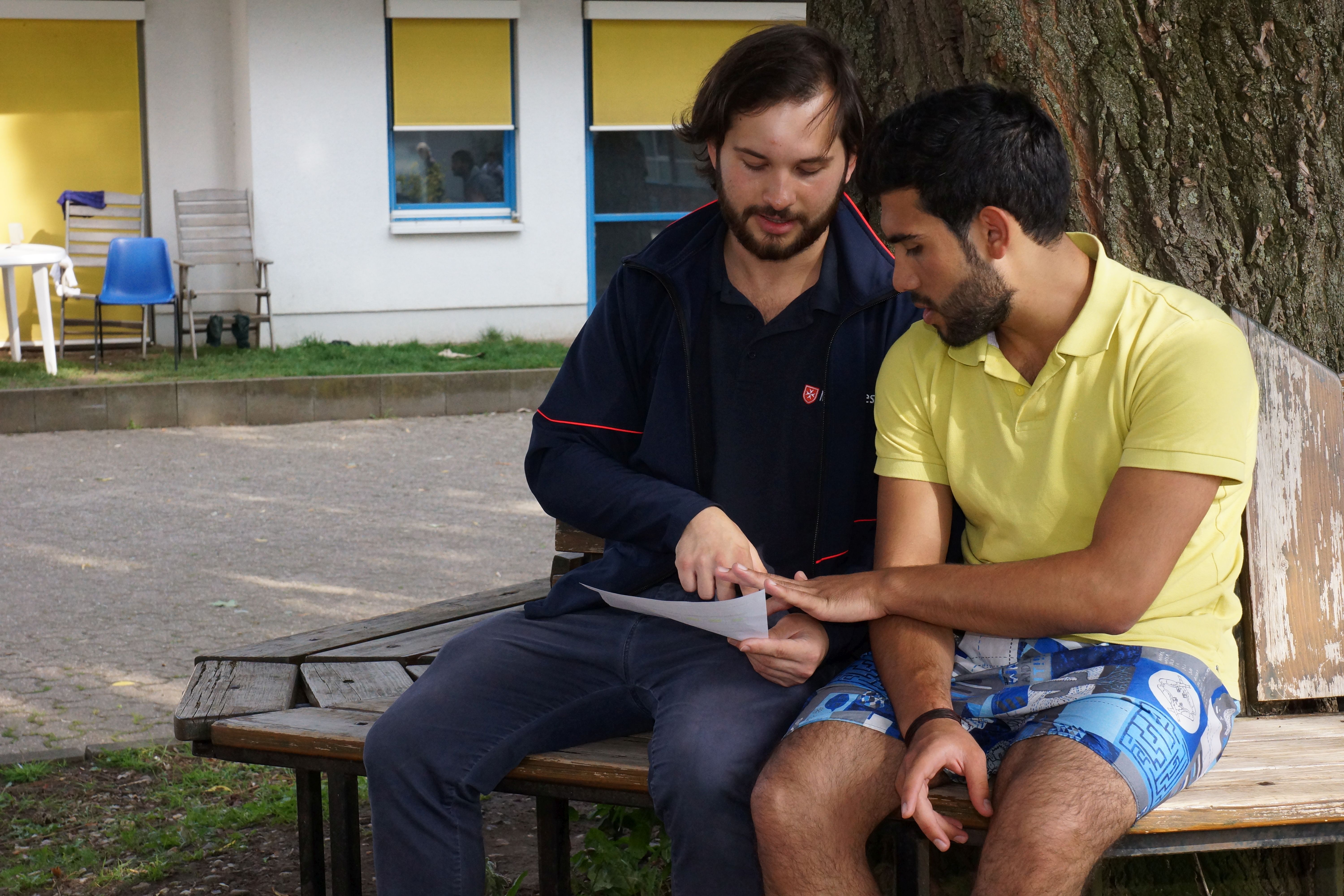 Zwei Männer sitzen auf einer Bank und schauen gemeinsam auf ein Blatt Papier.