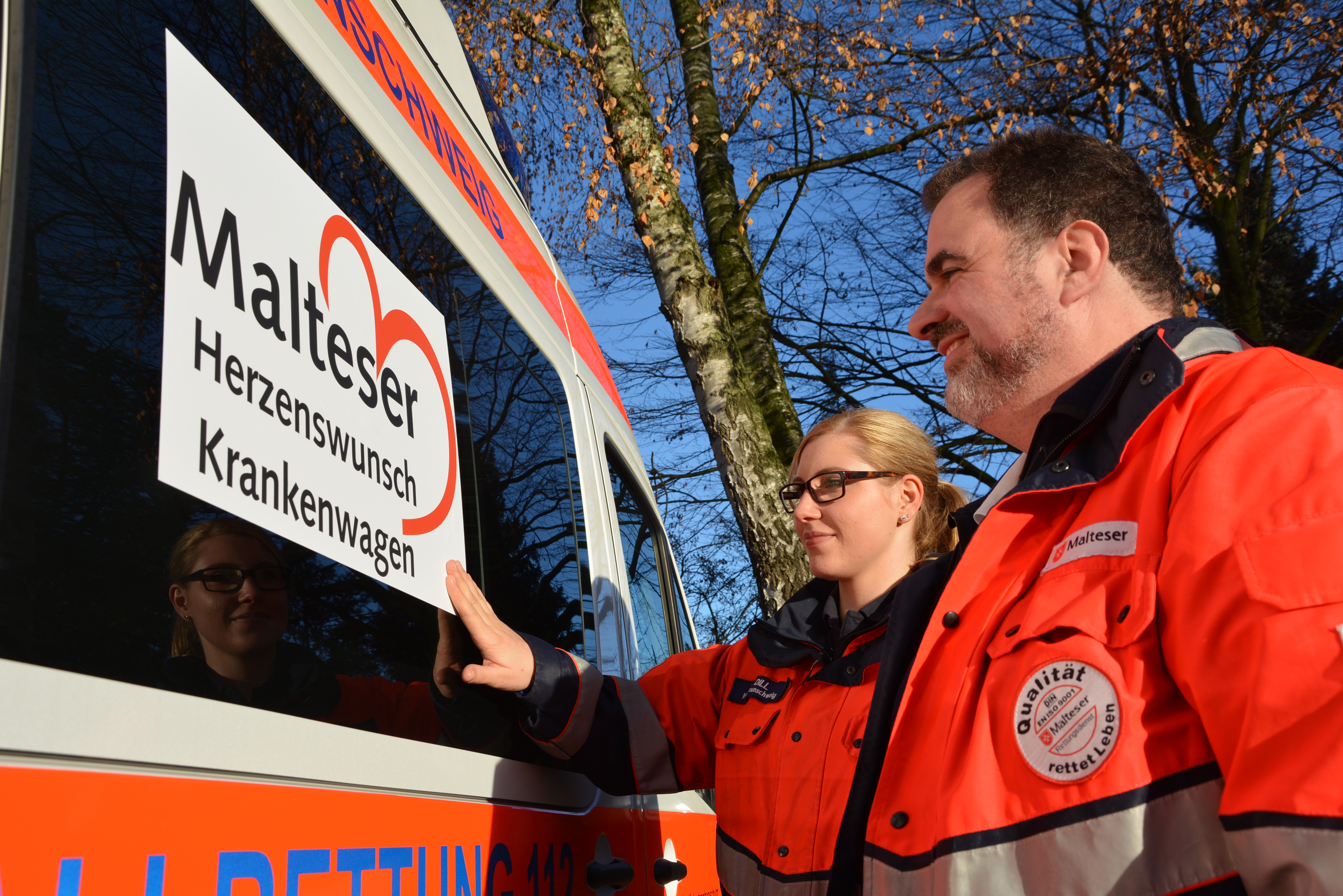 Zwei Mitarbeitende der Malteser stehen vor dem Herzenswunsch-Krankenwagen und schauen sich das Logo auf dem Auto an.