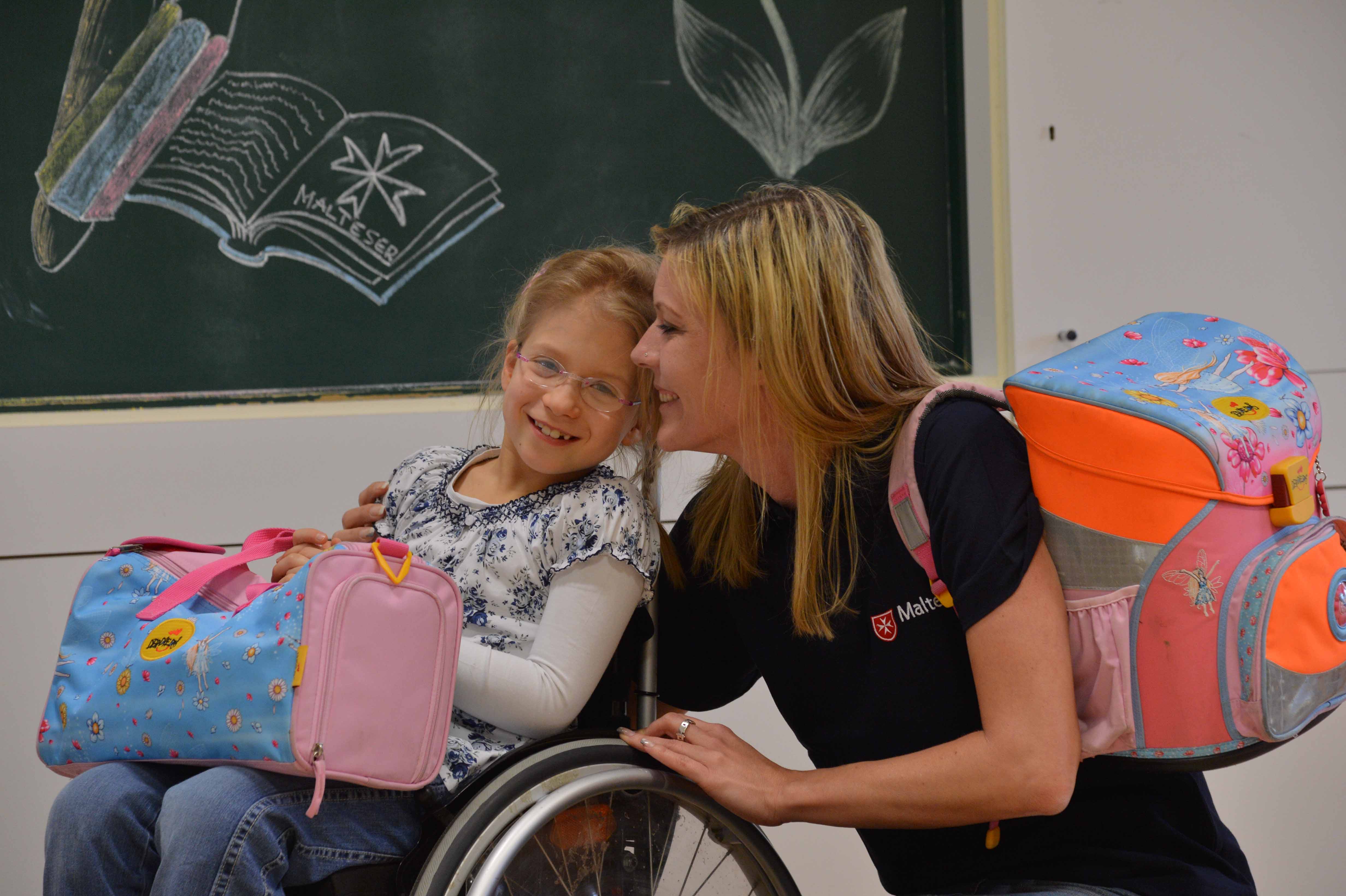 Eine Schulbegleiterin der Malteser sitzt neben einem Mädchen im Rollstuhl im Klassenzimmer.