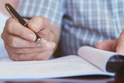 Eine ältere Hand hält einen Stift und unterzeichnet ein Blatt Papier.