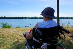 Eine ältere Frau sitzt im Rollstuhl und blickt auf einen See.