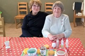Eine Ehrenamtliche und eine ältere Frau sitzen an einem Tisch vor Farben und Keramik.