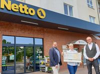 Zwei Männer und eine Frau stehen vor einer Netto-Filiale in Magdeburg. Die Frau hält einen großen Spendenscheck ins Bild.